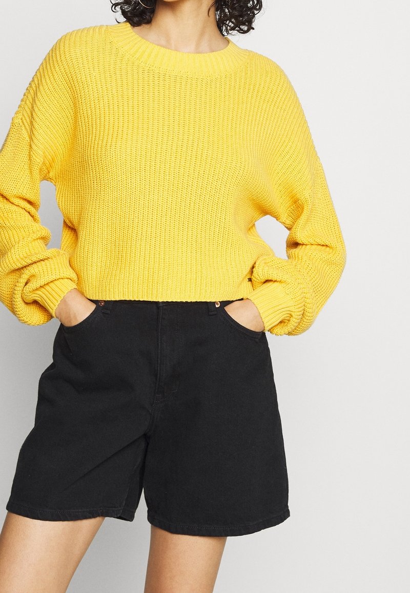 Suéter amarillo de punto con textura acanalada y hombros caídos, combinado con pantalones cortos de mezclilla negros de tiro alto con bolsillos laterales.
