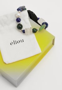 Pulsera de cuentas multicolores que presenta varias piedras, incluyendo verde, azul y blanco. Presentada en una bolsa de tela blanca sobre una caja degradada.
