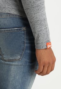 Harmaa pitkähihainen paita, pehmeästi teksturoitu, yhdistetty sinisiin farkkuihin, joissa näkyy takatasku ja oranssi merkkilappu.