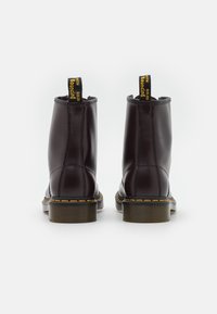 Un par de botas de cuero marrón oscuro hasta el tobillo con costuras amarillas, lengüetas de tira y suelas de goma gruesas, vistas desde atrás sobre un fondo blanco.