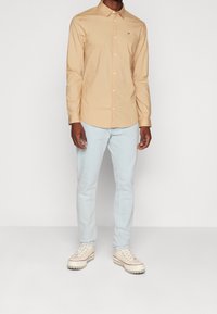 Hombre con camisa de botones beige, jeans azul claro y zapatillas blancas con cordones de color crema, de pie contra un fondo blanco.