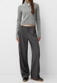 Szary prążkowany sweter z wysokim kołnierzem, zestawiony z ciemnoszarymi spodniami o szerokich nogawkach oraz czarnymi butami z brązowymi podeszwami, na białym tle.