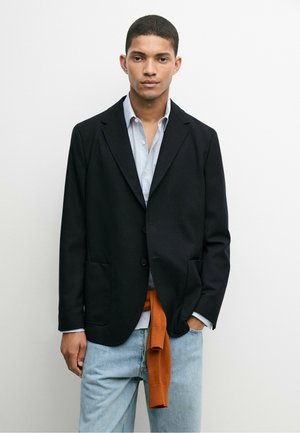 Ung mand iført en sort blazer over en lyseblå skjorte, blå jeans og en orange sweater bundet om livet, stående mod en hvid væg.