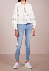 Blusa blanca bordada con un cuello atado, que presenta patrones florales. Pantalones vaqueros ajustados de color azul claro con rayas blancas en los laterales y dobladillos deshilachados. Zapatillas blancas.