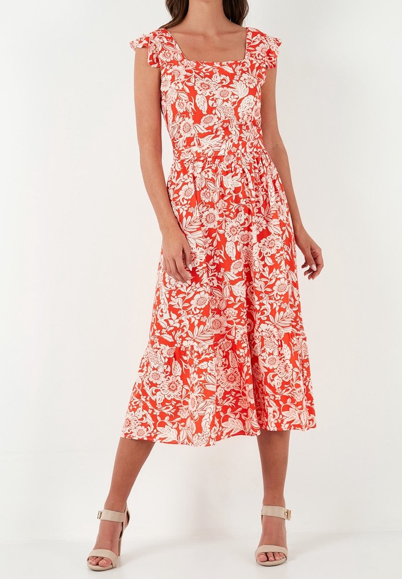 Femme portant une robe midi orange sans manches avec un col carré, imprimé floral blanc, et des talons beige à bride autour de la cheville, sur fond blanc.