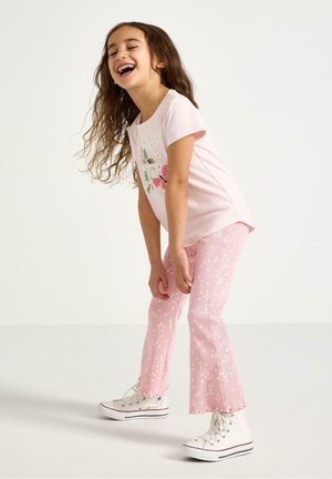 Ung pige, der griner, iført en lyserød blomster-T-shirt og matchende lyserøde blomsterbukser samt hvide høje sneakers, stående mod en ensfarvet baggrund.