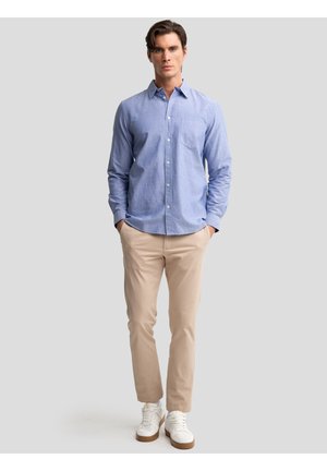 Mees seisab käed taskutes, kandes helehalli nööbitavat särki, beeže pükse ja valgeid tenniseid ühtlase taustaga.