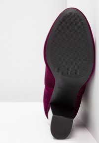 Zapato de terciopelo burdeos con tacón afilado y suela de goma negra texturizada. Parte superior lisa con una forma elegante y estilizada.