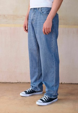 Isik, kes kannab lahtiseid siniseid teksapükse ja must-valgeid tossusid, seisab siseruumis kahvatu seina vastu.