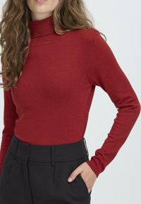 Sarkans, pieguļošs augstais kakls džemperis, izgatavots no mīksta materiāla ar garām piedurknēm, savienots ar augsto jostasvietu melnajām biksēm ar rāvējslēdzēju.