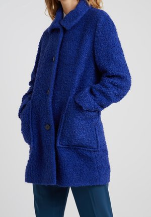 Kobieta w niebieskim, o długości do połowy uda, fakturowanym płaszczu z guzikami, dużymi przednimi kieszeniami i kołnierzem, z rękami włożonymi do kieszeni.