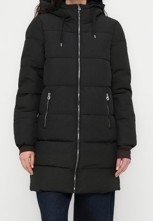 Femme portant un long manteau d'hiver noir matelassé avec capuche et fermeture éclair à l'avant, debout devant un fond blanc uni.