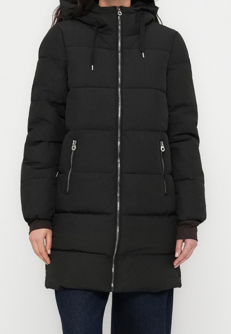 Femme portant un long manteau d'hiver noir matelassé avec capuche et fermeture éclair à l'avant, debout devant un fond blanc uni.