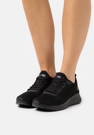 Jambes portant des chaussures de sport Skechers noires avec tiges en mesh à lacets et semelles rembourrées sur fond blanc.