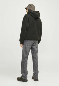 Isik, kes kannab musta fliisist kapuutsi, halli teksapükse, musti jalanõusid ja oliivrohelist kaabut, seisab seljaga vaataja poole tasasel taustal.