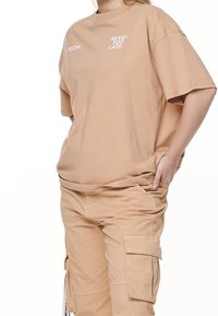 Béžové oversized tričko z bavlny s krátkými rukávy, s bílým nápisem "NEVER TOO LATE" a "PGDR" na přední straně. Nosené s odpovídajícími kalhotami.