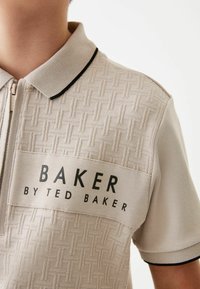 Polo beige con motivo tessuto testurizzato, chiusura a zip, profilo del colletto nero e logo "BAKER BY TED BAKER" sul petto.
