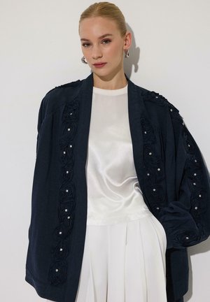 Jeune femme aux cheveux plaqués en arrière portant un haut en satin blanc et un pantalon à jambes larges plissé sous une veste noire ample avec une broderie florale texturée.