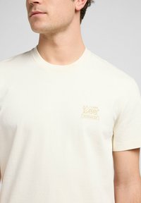 Kræmfarvet kortærmet t-shirt lavet af blødt stof, der har et lille beige broderet logo på venstre brystområde.