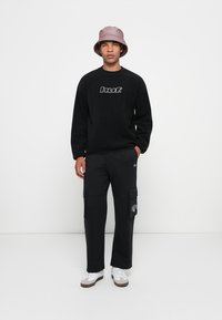 Ung mand iført en sort sweatshirt med "huf." logo, sorte cargo bukser, hvide sneakers og en mauve bucket hat, der står op ad en hvid væg.