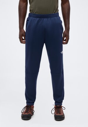 Homme portant un pantalon de survêtement bleu marine North Face et des chaussures de randonnée marron, debout devant un fond uni.