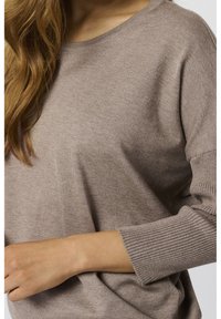 Femme portant un pull en maille taupe à manches longues avec des poignets côtelés et un col arrondi, son bras gauche plié sur sa taille.