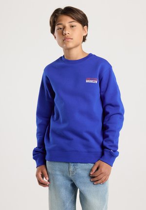 Un adolescent portant un sweat-shirt bleu avec le texte "Brooklyn" sur la poitrine et un jean bleu clair, se tenant devant un fond blanc uni.