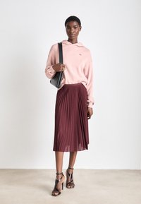 Sudadera rosa claro con un bolsillo frontal, combinada con una falda burdeos plisada y tacones altos negros con tiras. Accesorio de bolso de hombro negro.