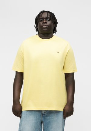 Άνδρας με σκούρο δέρμα και κοντά dreadlocks που φοράει ανοιχτό κίτρινο κοντομάνικο πουκάμισο και ανοιχτό μπλε τζιν μπροστά σε απλό λευκό φόντο.