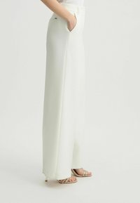 Witte wijde broek met een gladde textuur, met zijzakken en een subtiele metalen accenten op de tailleband. Gekoppeld aan beige sandalen met bandjes.