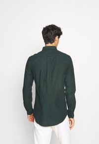 Camicia a maniche lunghe verde scuro in tessuto texturizzato, con colletto a camicia e piega sul retro, polsini aderenti e orlo curvo.