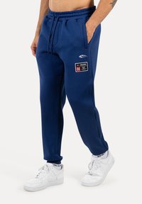 Pantaloni da jogging blu navy con vita elasticizzata, coulisse, tasche laterali e una toppa rettangolare con logo. Abbinati a sneakers bianche.