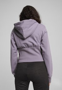 Felpa corta lilla con cappuccio, cintura elastica e tessuto testurizzato. Abbinate a leggings neri con motivo, che mostrano un design elegante.