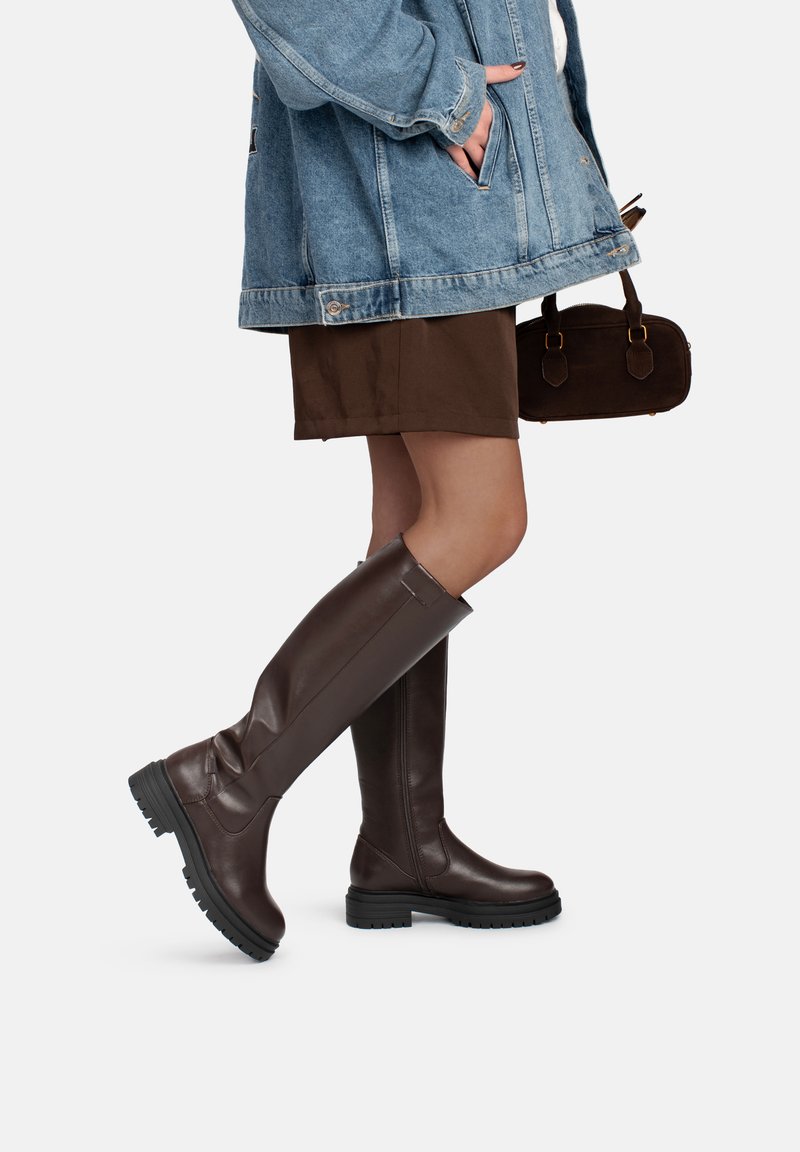 Bottes marron montant jusqu'au genou en cuir lisse, semelle en caoutchouc noire texturée, avec des détails minimalistes, associées à une veste en jean et une jupe marron.