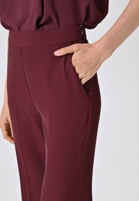 Bordeauxfarbene Hose mit glatter Textur, breitem Bund und Seitentasche. Der Stoff wirkt leicht und ist auf Komfort zugeschnitten.