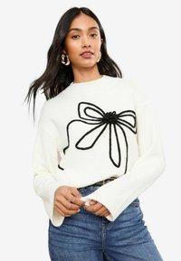 Weißer Pullover mit langen Ärmeln, der ein großes schwarzes Schleifenmuster auf der Vorderseite aufweist. Der Stoff wirkt weich und strukturiert. Sichtbare blaue Jeans.