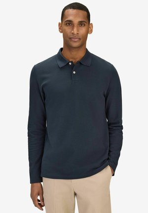 Man som bär en långärmad marinblå pikétröja och beige byxor, står med ena handen i fickan mot en vit bakgrund.