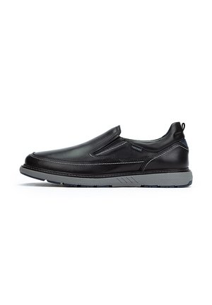 Zapato negro de cuero tipo slip-on con suela de goma gris, puntera redonda, paneles laterales elásticos y detalle de costura blanca en contraste.
