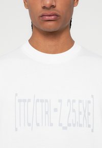 Λευκή κοντομάνικη μπλούζα από βαμβάκι, με γκρι κείμενο "[TTC/CTRL-Z_25.EXE]" εκτυπωμένο κεντρικά στην μπροστινή πλευρά.