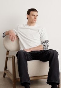 T-shirt blanc à manches courtes avec logo noir, superposé à une chemise à manches longues rayée noir et blanc. Jean foncé et assis sur une chaise claire.