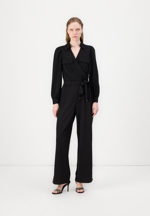 Schwarzer Jumpsuit mit Wickeldesign, langen Puffärmeln, zwei Brusttaschen, tailliertem Gürtel und weiter Beinform. Getragen mit schicken Riemchen-High Heels.