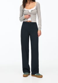 Cardigan gris à manches longues sur un débardeur blanc, associé à un pantalon large bleu marine foncé. Les chaussures sont vert foncé avec des semelles brunes.