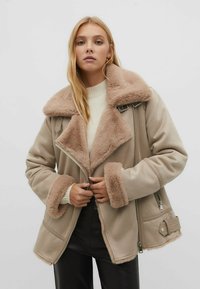 Beige Jacke mit Schaffellfutter, breitem Kragen, Reißverschluss und seitlichen Taschen. Weiche Textur und schichtartige Stoffdetails.