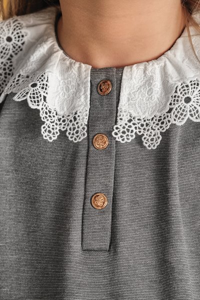 Grijze stof met een witte kant kraag met bloemmotieven. Drie bronzen knopen siëren de voorkant en voegen detail toe aan het ontwerp.