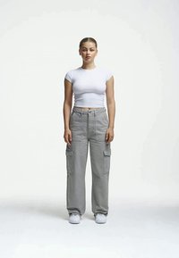 Tricou scurt, alb, asociat cu pantaloni cargo gri, lejeri, care au buzunare laterale. Modelul stă cu o expresie neutră pe un fundal simplu.