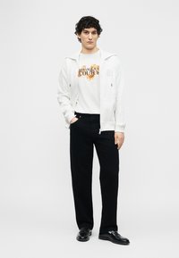 Νέος άνδρας με σγουρά μαλλιά που φοράει λευκό φούτερ με γραφικό σχέδιο, λευκό φούτερ με φερμουάρ, μαύρο παντελόνι και μαύρα δερμάτινα παπούτσια, όρθιος με το ένα χέρι στη τσέπη.