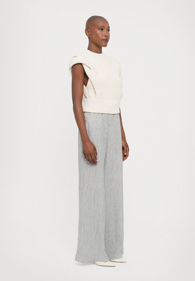 Beige knitted sleeveless top with textured design and raised shoulder detail, paired with light gray wide-leg striped trousers and white shoes.