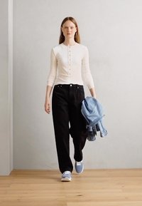 Haut beige clair à manches longues avec détail de boutons, pantalon noir à jambes larges et baskets bleues ; tenant une veste en denim clair. Design simple, look décontracté.