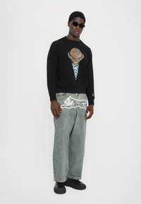 Zwarte sweatshirt met een graphic van een ijsco, gecombineerd met lichtblauwe denim broek met een cartoonkat-ontwerp op de achterzak.