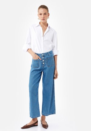 Camisa blanca ligera con mangas enrolladas, combinada con pantalones de denim de tiro alto en azul y pernera amplia, y zapatos planos marrones. Presenta bolsillos frontales y detalles de botones.